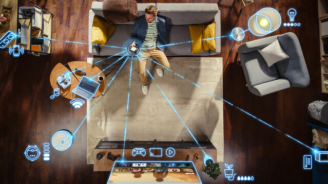 Top View Of Caucasian Man In the Loft Apartment Sitting Down on The Couch and Connecting Smartphone to Convenient Smart Home System. VFX Edit Visualizing Connected Devices. Laptop, TV, Speaker.