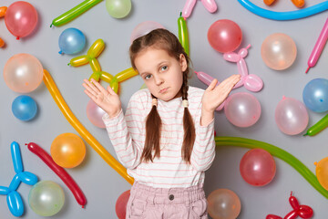 Fototapeta premium Uncertain confused little girl with pigtails standing against gray wall with balloons decoration shrugging shoulders looking at camera with puzzled facial expression.