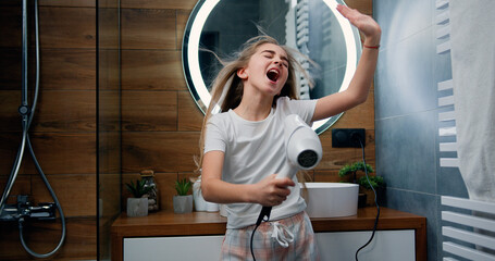Beautiful blond little girl in bathrobe singing a song and dancing with a hair dryer. Child sings and dances in a shower after taking shower in bathroom. - obrazy, fototapety, plakaty