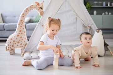 a blonde girl in a bright children's room plays wooden cubes with her younger sister at the wigwam and rejoices and smiles © Any Grant