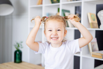 Fototapeta premium a little funny baby blonde girl in a bright library at home indulges in ponytails and smiles