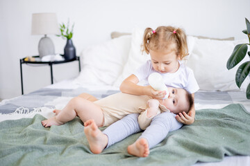 sisters are feeding baby milk from a bottle on the bed at home, the older sister is holding the baby at home on the bed, the concept of baby food