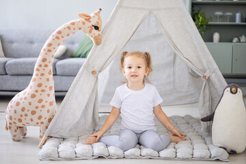 a blonde girl in a bright children's room is playing wooden cubes with a giraffe at the wigwam and is happy and smiling © Any Grant