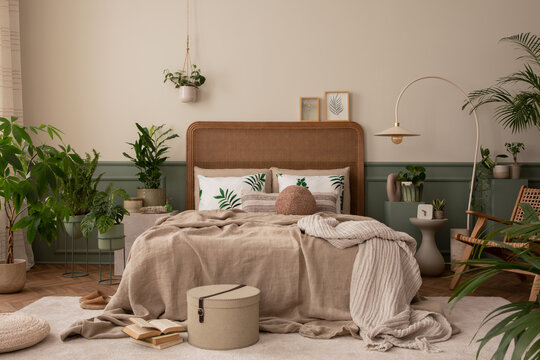 Creative composition of bedroom interior with cozy bed, plants, beige beding, stylish lamp, rattan armchair, pouf, carpet, brown slippers, wooden stool and personal accessories. Home decor. Template.