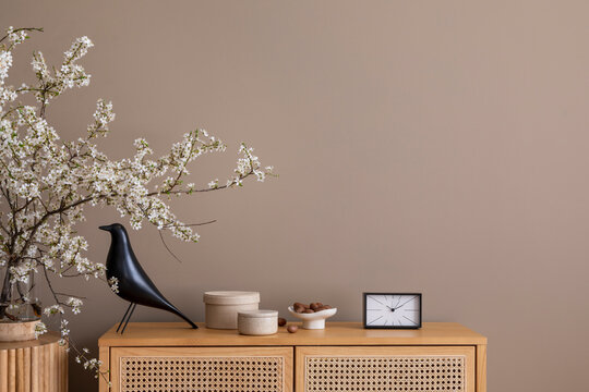 Minimalist composition of spring living room interior with copy space, rattan sideboard, blooming branch, stylish sculpture, clock, round box and personal accessories. Home decor. Template.