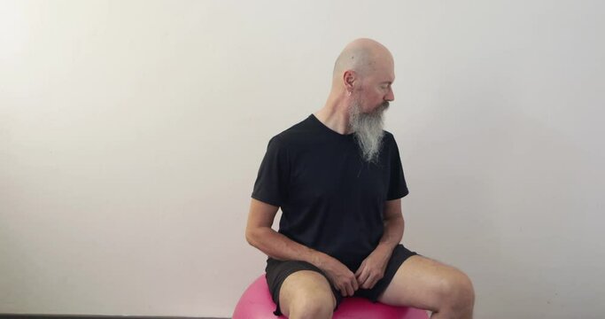 50 year old mid adult man with long beard sitting on fitness ball, doing neck and upper spine exercise at home moving head at side upwards and downwards 