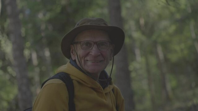Senior man enjoys hiking and exploring flora and fauna in deep forest. Old hiker wearing hat walks in forest with delighted expression