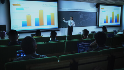 Young University Professor Explaining the Importance of Branding and Marketing Campaigns to a Group of Diverse Multiethnic Students in a Dark Auditorium. Female Teacher Showing Business Charts Data