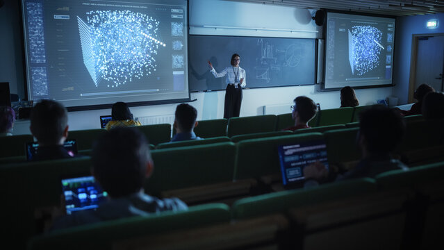 Young University Professor Explaining the Importance of Artificial Intelligence to a Group of Diverse Students in a Dark Auditorium. Female Teacher Showing Neural Network Eco System on Two Big Screens