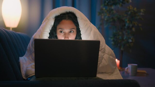 Hispanic freelancer girl Latino woman Indian Arabian female at night evening home at couch sofa under blanket cover duvet working with laptop watching movie at computer browsing social media addict