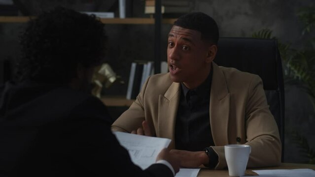 African American businessman chastises his worker for inefficient work. A black man in a business suit sits at a table and explains something to his worker.