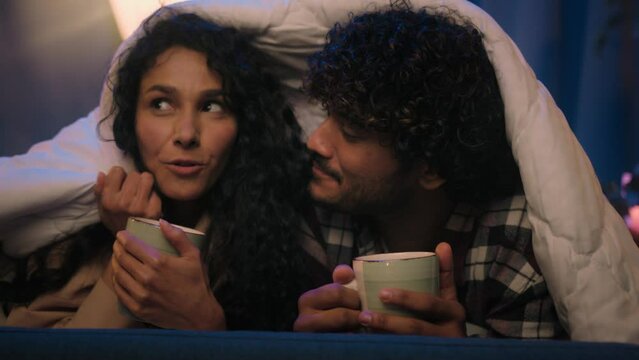 Multiracial couple love diverse boyfriend and girlfriend man and woman drink tea cups talking laughing cuddling affectionate romantic bonding under warm blanket cover duvet at cozy evening night home