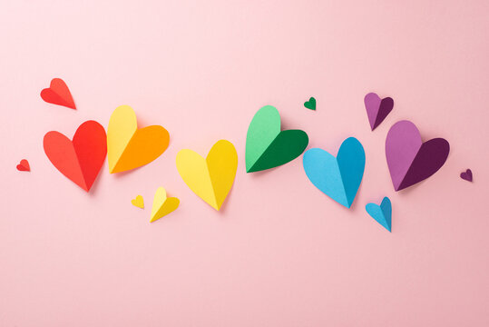 A symbolic flat lay of LGBTQ History Month parade accessories. Top view of rainbow flag colored paper hearts, arranged on a pastel pink backdrop