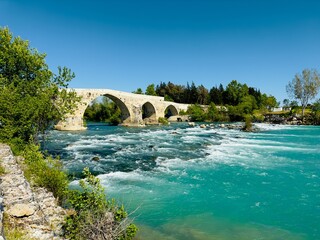 Flowing river beneath the historic Aspendos bridge, surrounded by lush vegetation and the timeless...