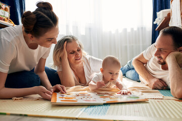 Fototapeta premium Parents together with their child play and study a children's book, lying on the floor in a bright children's room. Happy friendly family concept.