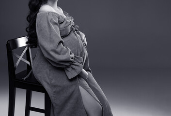 A pregnant woman in a dress is sitting on a chair on a gray background. The belly of a pregnant woman. Studio pregnancy photo shoot. Black and white photo. Copy space.