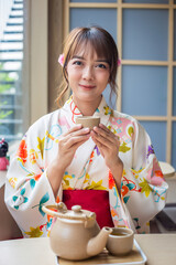 young woman wearing a traditional kimono sits in a Japanese style house raising a teacup to drink,...