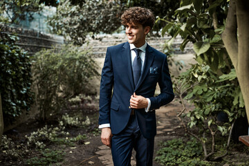 Attractive young man wear dark blue suit posing in green garden