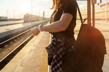 Female person standing on railway station in evening and looking at watch on her hand, train on background.