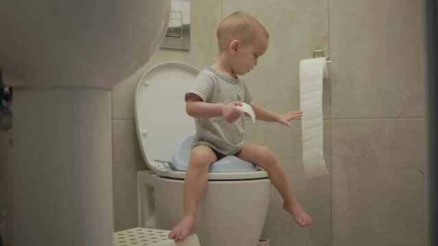 baby toddler sitting on toilet learning to clean his hygiene bottom butt anus toilet paper. potty training child in bathroom. little boy kid pulls toilet paper sweep his back after poop poo defecate