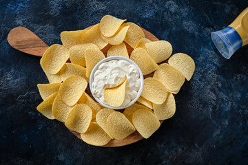 Top view potato chips, sauce in a white bowl on a wooden cutting board.