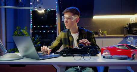 Teenager boy according to a video he watches on his laptop and tighten the nut with a screwdriver to assemble car toy, creating electronic AI technology workshop school lesson. Hobbies for advanced