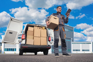 Delivery man with box. Courier near minivan. Man from transport company is holding tablet. Courier picks up parcels from warehouse. Delivery man near industrial buildings. Logistics company employee
