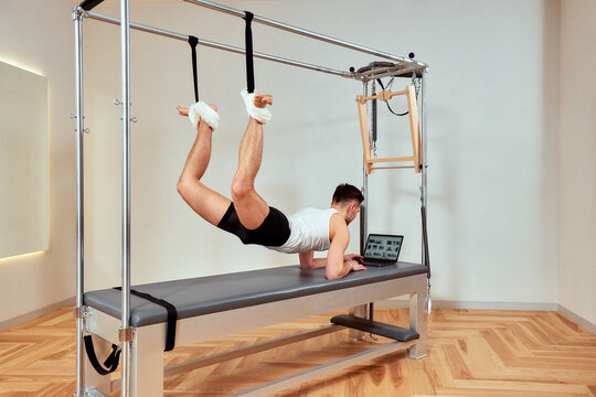 Young man doing pilates, stretching his body on a reformer and looking at a laptop, online program classes, training for muscles and mind