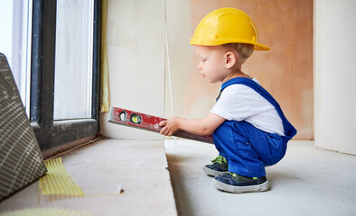 Child checking wall surface with spirit level in apartment. Kid in work overalls and safety...
