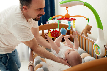 A happy dad puts his baby in a crib with a musical toy. The father hugs his baby. Happy childhood.