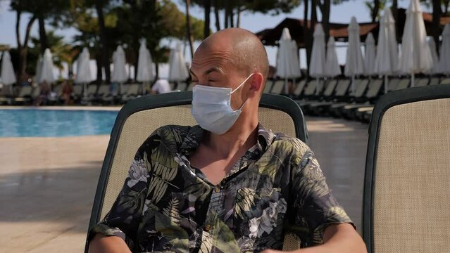 Portrait of a young sick man with symptoms of coronavirus in a hotel on vacation, he is sitting on a chaise longue by the pool and coughing. A sick man on vacation in a hotel.