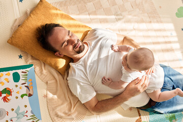 Happy dad holds his newborn baby in his arms while lying on the floor in a bright child's room. The...