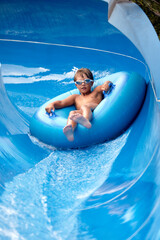 Joyful boy in swimming goggles slides down the blue water slide on an inflatable boat in the water...