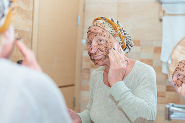 Mature woman standing at the mirror in the bathroom and putting a scrub on her face. Elderly woman taking care of her face