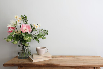 Fototapeta na wymiar Moody spring still life. Wooden bench, table composition with cup of coffee, tea and old books. Beautiful floral bouquet with white, pink tulips, daffodils. Hawthorn, green guelder rose flowers. Wall.