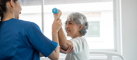 Fototapeta premium Young physiotherapist helping senior mature asian woman grey hair work out with dumbbells, to recover from injury at health centre in physical therapy session.