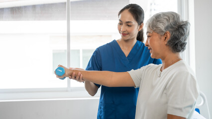 Fototapeta premium Young physiotherapist helping senior mature asian woman grey hair work out with dumbbells, to recover from injury at health centre in physical therapy session.
