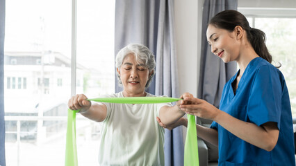 Obraz premium Young physical therapist caregiver assisting mature asian woman grey hair doing exercise with elastic bands at physiotherapy clinic.