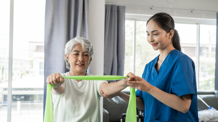 Fototapeta premium Young physical therapist caregiver assisting mature asian woman grey hair doing exercise with elastic bands at physiotherapy clinic.