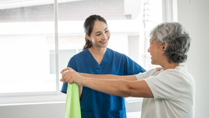 Obraz premium Young physical therapist caregiver assisting mature asian woman grey hair doing exercise with elastic bands at physiotherapy clinic.
