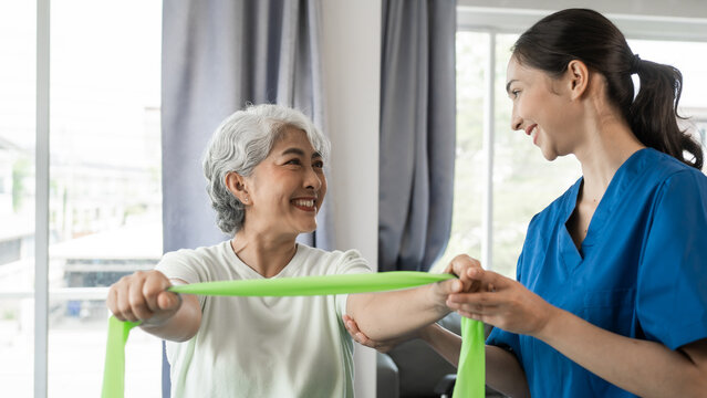 Young physical therapist caregiver assisting mature asian woman grey hair doing exercise with elastic bands at physiotherapy clinic.