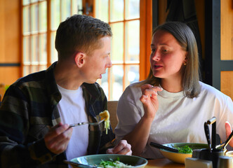 Sitting by a bright window in a cozy restaurant, a couple shares a meal together during lunchtime. The natural light illuminates the space, they chat and enjoy their food.