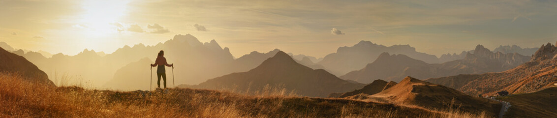 Panorama of  sunset mountains and a woman standing with trekking poles. - 601456056