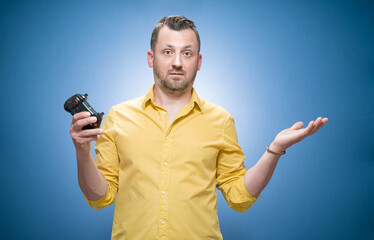 Man shrugs shoulders with game controller - joystick uncertainty, cant make decision looking clueless at camera over blue background, dresses in yellow shirt