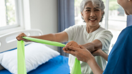 Young physical therapist caregiver assisting mature asian woman grey hair doing exercise with elastic bands at physiotherapy clinic.