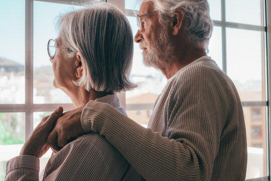 Melancholic senior family couple of Caucasian people hugging each other with love, elderly man and woman with glasses support each other in a moment of sadness looking out the window