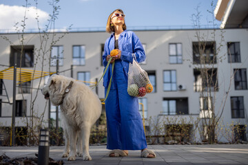 Young woman in blue pajamas walks with her dog and carry mesh bag full of fresh fruits and...