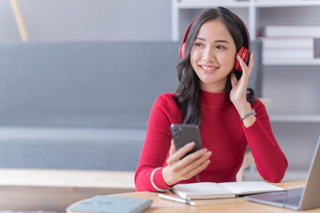 Asian beautiful woman in the living room,listening to music and playing social media with mobile phone and laptop for relaxing, studying online. University student concept.