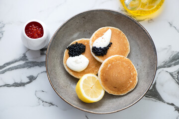 Pancakes served with black caviar and sour-cream in a grey plate, horizontal shot on a white marble background, elevated view