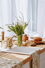 The interior of a stylish kitchen with a wooden worktop on which there are kitchen utensils, a towel and a vase with white tulips. Concept of good morning and breakfast at home.